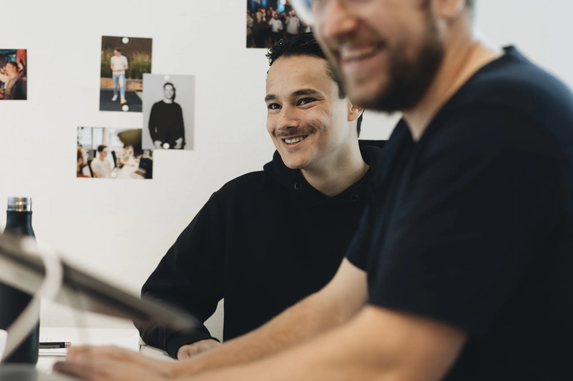 Hier zien we Dani vrolijk kijken, hoogstwaarschijnlijk naar een beeldscherm. Achter Dani zijn er meerdere, kleine foto's van team Stimmt. Voor Dani zien we een man, glimlachend, waarvan niet het gehele gezicht te zien is, echter alleen zijn glimlach en baard.