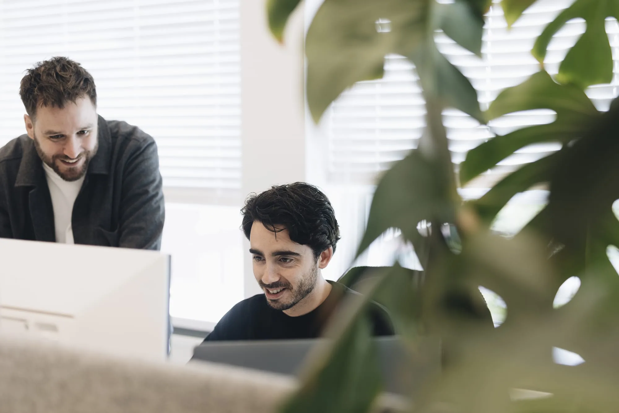 Bram is aan het werk achter zijn werklaptop en een collega staat naast hem. Lachend kijken ze naar het beeldscherm.